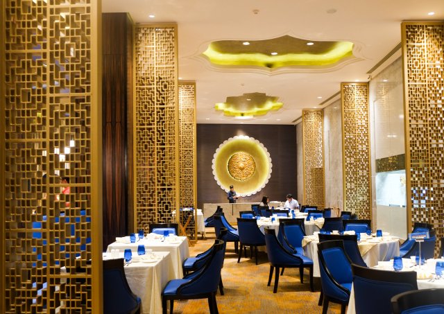 Gold lattice screens with bright blue velvet chairs at tables with white table clothes. Yellow clam shaped lighting.
