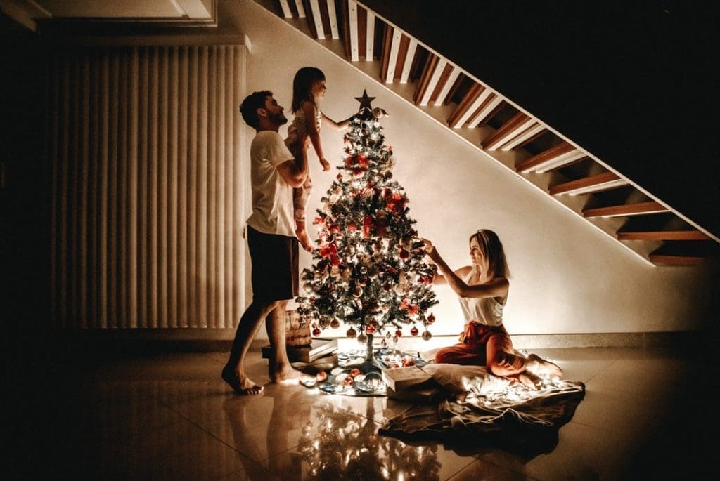 man lifting child next to christmas tree and women on floor putting decorations on tree