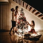 man lifting child next to christmas tree and women on floor putting decorations on tree
