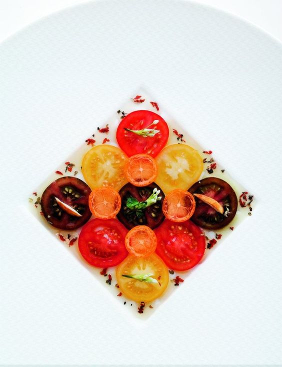 thinly sliced tomatoes in yello, red and deep red on a white cirular plate in a square shape.