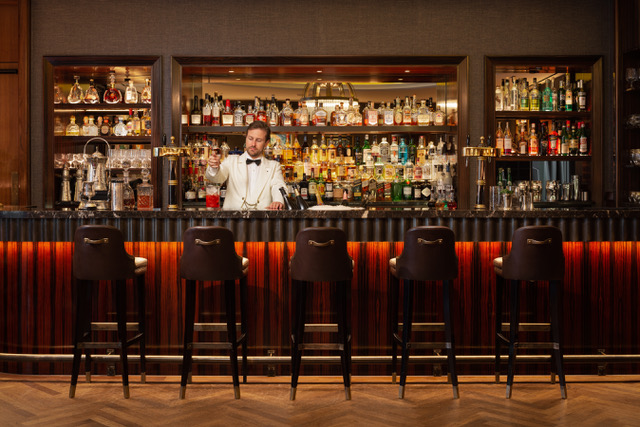 Red seats on wooden floor in front of spectacular bar packed with bottles and a barman in front in a white jacket