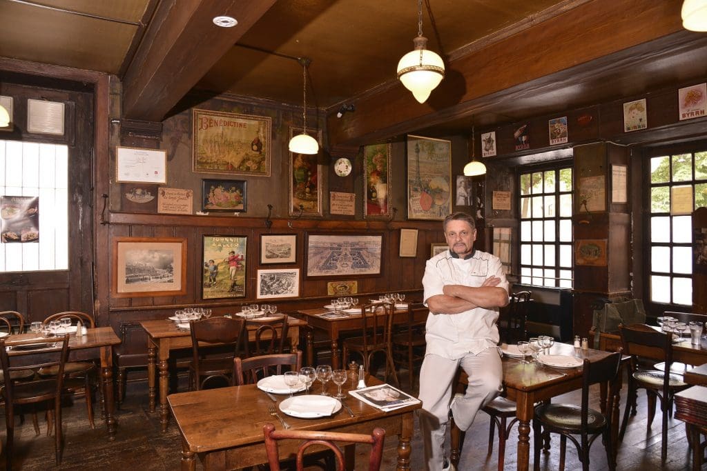 chef in white in an old style wooden walls, ceiling and floor restaurant with brown wooden tables and coloured small art works in the wall behind him