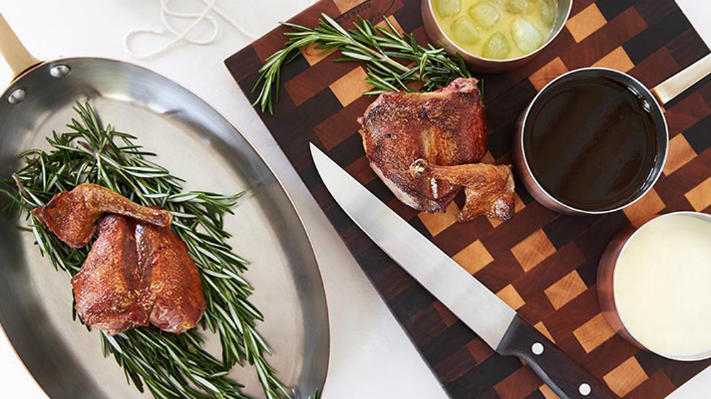 meat and rosemary on a chopping board with a knife, and meat and rosemary sprigs in a silver oval dish next to it.